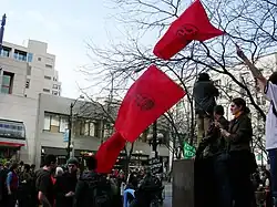 Three red flags with IWW logos being held above a crowd of people.