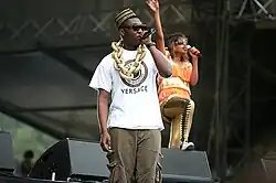 A man wearing an African-style hat, sunglasses, and a large gold chain, and holding a microphone. In the background a woman in brightly coloured clothing and sunglasses is also holding a microphone and is waving one hand in the air