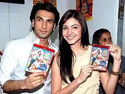 Anushka Sharma and Ranveer Singh holding up DVDs. While Sharma, wearing a pale yellow top is smiling directly at the camera, Singh, dressed in a white shirt, is looking away.