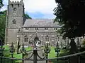Gray stone building with square tower at left hand end. Foreground includes grass area with gravestones, taken over the top of metal railings.