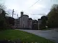 Stone building with slit windows and battlements. Foreground is road with grass verges.