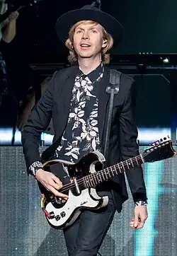 Musician Beck, wearing a black suit and hat, and holding an electric guitar.