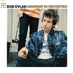 A photograph of Dylan seated in a blue jacket with a person standing behind him holding a camera