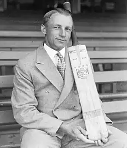 Man in double breasted suit, hair parted down the middle, sitting on a long bench in a sports stadium, posing with a cricket bat, held vertical and supported on his thigh.