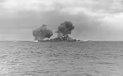 Black-and-white photograph of a warship firing the guns. Two columns of dark smoke drift from the ship