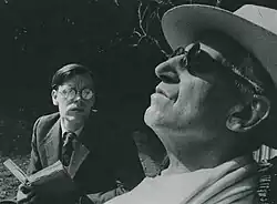 young bespectacled white man reading to an older, blind, man in a garden