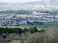 Multiple houses mostly with red roofs seen in a valley between the vegetation in the foreground and the hills beyond.