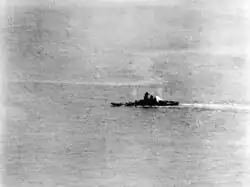 A large area of ocean with a warship in the middle distance. A plume of smoke is coming from the rear of the ship's superstructure, and the ship appears to be leaning to the left.