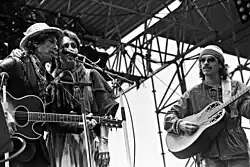 On stage outdoors, Dylan in a leather jacket and iconic porkpie hat & scarf, Baez wears a peasant skirt, sarape, tassled scarf around neck, her arm around Dylan's shoulder. Santana in workman's shirt & knit cap.