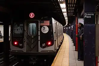 An E train, here composed of R160A cars is seen leaving the Queens Plaza station. The back of the train contains two red tail lights, a window on the left side, the American flag on the right side, and the MTA logo below the flag.