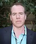 Bret Easton Ellis is pictured standing on front of a stone wall and greenery. He is wearing a blazer over a blue collared shirt and has a lanyard on his neck.