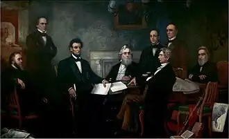 A dark-haired, bearded, middle-aged man holding documents is seated among seven other men