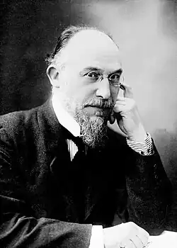 Middle-aged white man, balding, with neat moustache and beard, in formal day wear with wing collar and dark necktie. He is wearing pince-nez glasses.