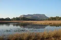 Daytime view of a small marshy lake surrounded by dry brush in winter. Behind the lake in the far-middle distance, a large stand of trees interposes itself in front of a lone massif in the far distance. The sun is out of view to the left.