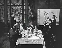 A black and white film still. A group of men sit around a dining table in the center. To the right, a man stands by and gestures at a large drawing of a dinosaur.