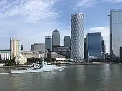 A gray naval patrol ship marked "P233" sails along a wide river in front of the Canary Wharf skyline, with modern glass skyscrapers and a partly cloudy blue sky in the background.