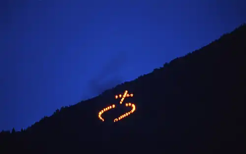 A photograph of a dusk scene. On the top, left, a dark blue triangle of dusk sky. On the bottom left, a black triangle of distant hillside. On the hillside, several fire points form a schematic round shape crowned by a Latin cross.