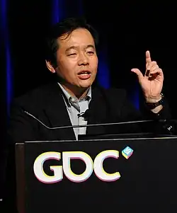 Portrait of a Japanese man wearing a black business jacket standing behind a podium with his left hand pointed upward.