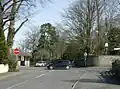 Street scene. Road junction with red stop sign. Two cars. The sides of the roads are stone walls with large trees showing above them.