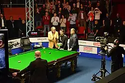 The audience applauds as Trump stands behind the trophy which is sat on the snooker table