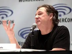 Man speaking into a microphone at a panel table, gesturing with one hand in front of a WonderCon backdrop