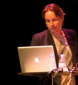 A 41-year-old man with light brown hair wearing a coat over a shirt and looking down at the screen of a laptop