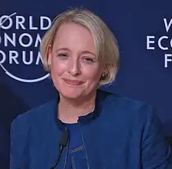 A woman with short blonde hair wearing a blue jacket speaks at a podium in front of a World Economic Forum backdrop