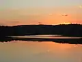 A red-orange sunset and the black silhouette of a line of trees are reflected in a smooth lake