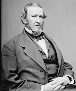 A man in his late forties with receding hair and a full beard. He is wearing a white shirt with a black vest and jacket and is facing right