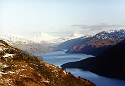 In the distance there are high snow-capped mountains, out of which a sinuous lake emerges. In the foreground lower brown hills surround the water.