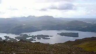 A view from above a lake containing several wooded islands with a wilderness of hills and moor beyond.