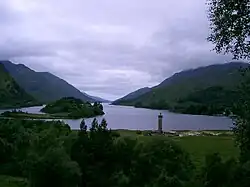 A gloomy, cloud-covered sky above a large lake surrounded by green hills. There is a wooded islet at left and a tall circular tower in the foreground topped by a statue.