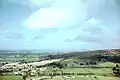 The houses and church of a small village can be seen in the bottom left of the picture. It is surrounded by a patchwork of fields with some trees on a hillside. Large hills in the distance.