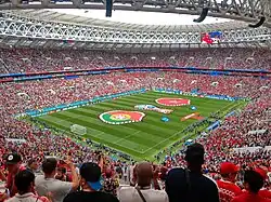 A football pitch is surrounded by a large audience of people mostly dressed in red.