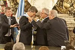 A smiling Macri puts on a blue-and-white sash with the help of Federico Pinedo and two other men