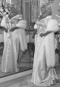Mae West posing in a glamorous dress with a confident smile, 1953.