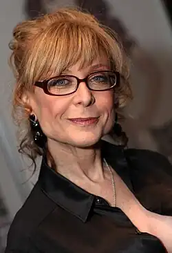 Portrait of a smiling woman with dark blonde hair, wearing thick-rimmed glasses and a black collared shirt