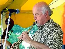 A man wearing a patterned dress shirt, performing a wind instrument while standing behind a microphone