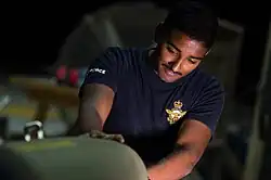 A man wearing a blue T-shirt with a military logo working on a green bomb