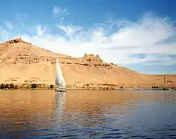 A photograph of a sailboat on a wide, calm, blue river. In the background is a blue sky with clouds and a rocky hill.