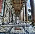 A colonnaded portico with a decorated ceiling and statues along the wall, overlooking the courtyard.