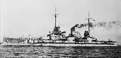 A large warship steams at low speed; gray smoke drifts from the two smoke stacks
