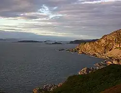 A sunlit rocky promontory at right and a calm, dark sea at left. Small dark islands pepper the waters in the distance