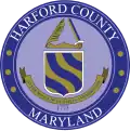 A round seal featuring a blue-and-gold shield and ribbon with a blue border