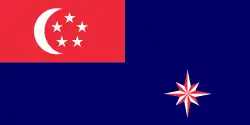 A red rectangle at the top left corner of the flag, charged with a white crescent and five white stars arranged in a pentagon. The rest of the flag is coloured blue. At the bottom right part of the flag is an eight pointed star that is alternating red and white.