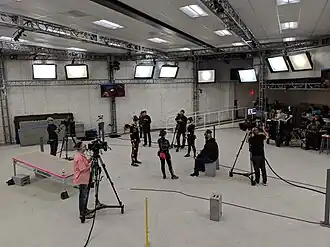 A bright white stage with actors in black suits and camera operators