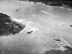 Black and white aerial photograph showing a body of water with a large warship near the shore