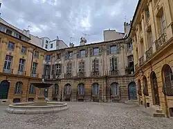 A cobbled courtyard surrounded by old yellow-stone buildings with shuttered windows and a central fountain under an overcast sky in Aix-en-Provence.