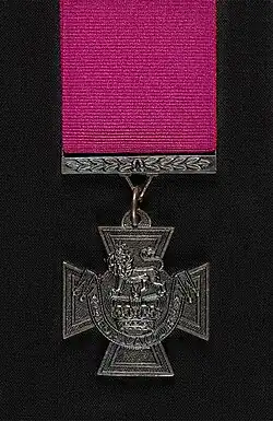 A bronze cross pattée bearing the crown of Saint Edward surmounted by a lion with the inscription "for valour". A crimson ribbon is attached