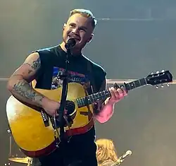 A photograph of Zach Bryan performing on stage with a guitar